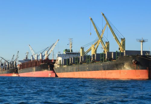 Four bulk carrier vessels lined up along a wharf, taking on cargo with their cranes.