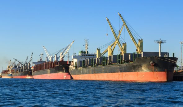 Four bulk carrier vessels lined up along a wharf, taking on cargo with their cranes.