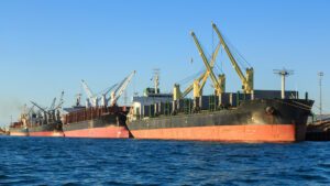 Four bulk carrier vessels lined up along a wharf, taking on cargo with their cranes.