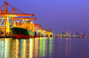 Industrial Container Cargo freight ship with working crane bridge in shipyard at dusk for Logistic Import Export with oil plant background