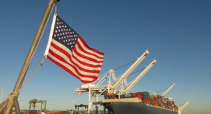 An American flag waves at a US port, where a cargo ship is docked beneath a giant crane on a blue sky day. The image symbolizes Made in America, Made in the USA, industry, global economy, pride, strength, power, patriotism, trade, manufacturing, and back to work.