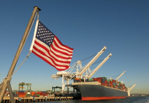 An American flag waves at a US port, where a cargo ship is docked beneath a giant crane on a blue sky day. The image symbolizes Made in America, Made in the USA, industry, global economy, pride, strength, power, patriotism, trade, manufacturing, and back to work.