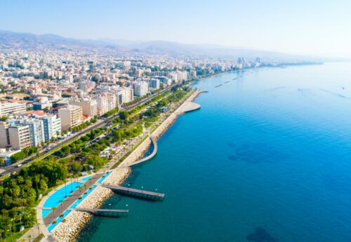 Aerial view of Molos Promenade park on the coast of Limassol city centre in Cyprus. Bird`s eye view of the jetties, beachfront walk path, palm trees, Mediterranean sea, piers, rocks, urban skyline and port from above.