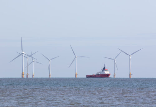 Renewable energy offshore wind farm turbines with passing maintenance supply vessel ship. Windfarm on the sea horizon with boat sailing between towers. Sustainable resource green power development